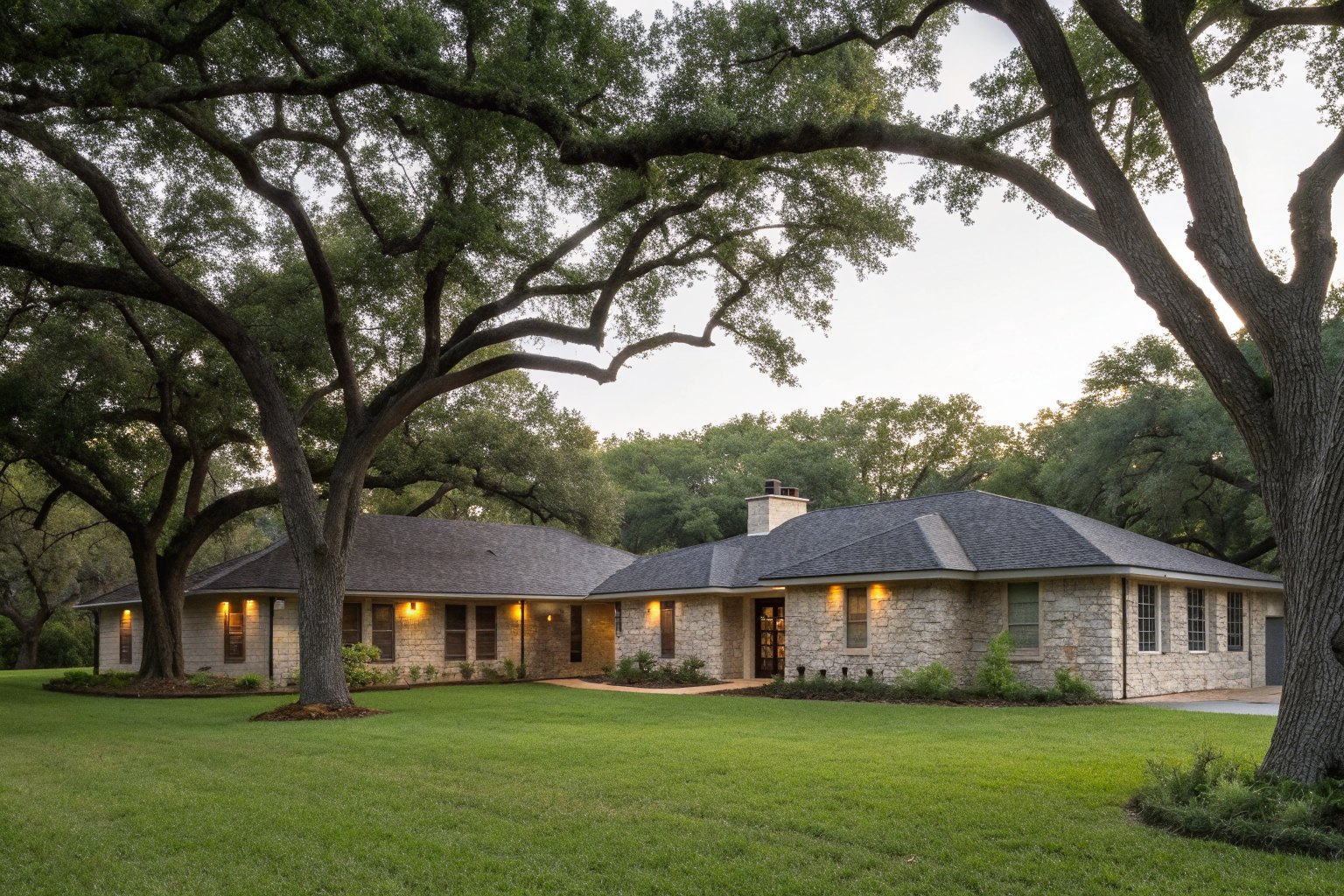 A one-story limestone and brick ranch house with a charcoal shingle roof under live oak trees